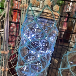May include: A glass jar with a blue and white string net around it. The jar has a silver lid and contains white fairy lights.