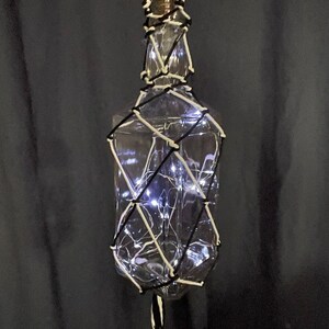 May include: A clear glass bottle wrapped in white and black string with white LED lights inside. The bottle is hanging from a black and white cord.