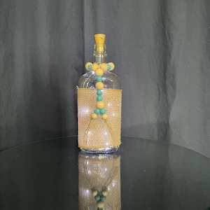 May include: A clear glass bottle with a burlap wrap and a string of wooden and turquoise beads. The bottle is lit from within with white lights.