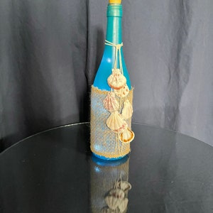 May include: A blue glass bottle with a cork top, decorated with burlap and seashells. The bottle is sitting on a black surface.