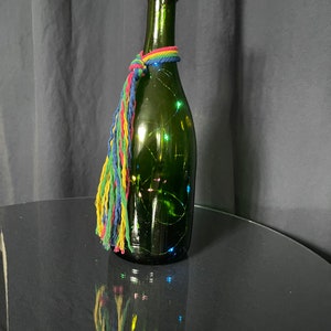 May include: A green glass bottle with a cork top and a colorful tassel. The bottle is filled with white fairy lights.