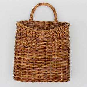 May include: A rectangular woven wicker basket with a rounded handle. The basket is a warm brown color and has a textured surface. The handle is also woven and is attached to the top of the basket.