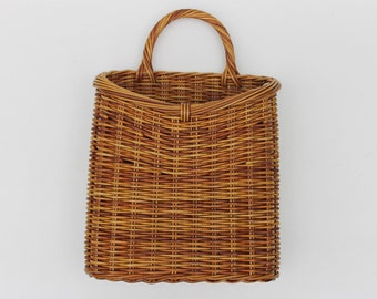 May include: A rectangular woven wicker basket with a rounded handle. The basket is a warm brown color and has a textured surface. The handle is also woven and is attached to the top of the basket.