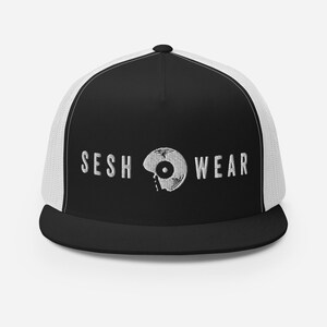 Op de afbeelding: Zwarte en witte truckercap met een wit mesh achterpaneel. De voorkant van de cap heeft een wit geborduurd ontwerp van een plaat met de tekst "SESH WEAR" eronder.