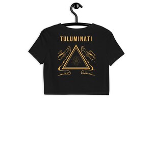 Puede incluir: Camiseta negra de corte corto con un gr&aacute;fico dorado de un tri&aacute;ngulo con un ojo en su interior, rodeado de dos manos. El texto "TULUMINATI" est&aacute; por encima del gr&aacute;fico.