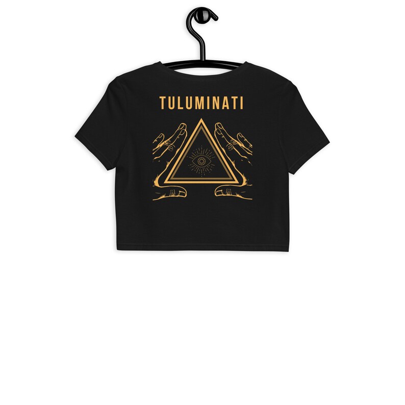 Puede incluir: Camiseta negra de corte corto con un gr&aacute;fico dorado de un tri&aacute;ngulo con un ojo en su interior, rodeado de dos manos. El texto "TULUMINATI" est&aacute; por encima del gr&aacute;fico.