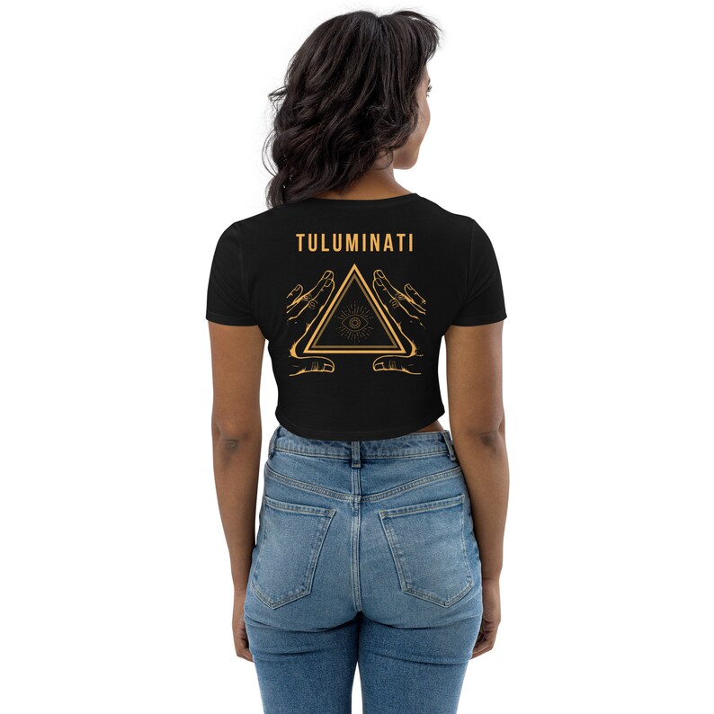 Puede incluir: Camiseta negra de corte corto con un gr&aacute;fico dorado de un tri&aacute;ngulo con un ojo en su interior y dos manos a cada lado. El texto "TULUMINATI" est&aacute; por encima del gr&aacute;fico.