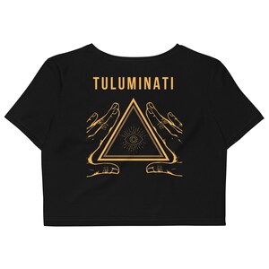 Puede incluir: Crop top negro con un gr&aacute;fico dorado de un tri&aacute;ngulo con un ojo dentro, rodeado de dos manos. El texto "TULUMINATI" est&aacute; por encima del gr&aacute;fico.