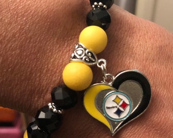 May include: A black and yellow beaded bracelet with a silver heart charm featuring a Pittsburgh Steelers logo.