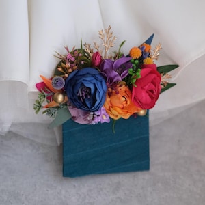 May include: A teal blue velvet box with a colorful floral arrangement. The flowers are red, orange, yellow, blue, purple, and green.
