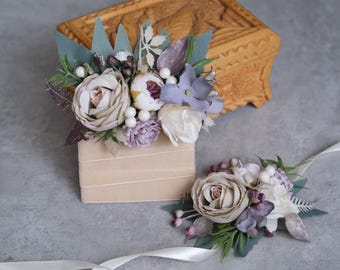 Op de afbeelding: Een set van twee bloemsieraden met witte en grijze rozen, paarse en blauwe bloemen en groene bladeren. De bloemsieraden zijn vastgebonden met een wit lint.