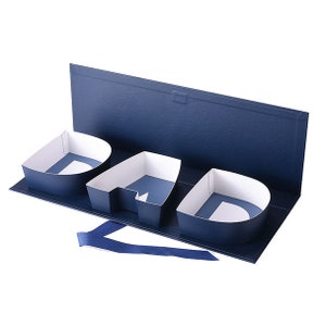 May include: A navy blue gift box with the word "DAD" formed by three separate compartments. The interior of each letter is white, with a blue base. A blue ribbon is attached to the box.