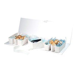 May include: A white gift box with an open lid, revealing several small white containers. Some containers hold gold-wrapped candies nestled in blue tissue paper. Other containers hold black and white items.