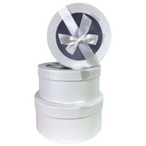 May include: Three white round gift boxes stacked on top of each other. The top box has a clear window and is adorned with a white satin ribbon tied in a bow. The boxes are a clean, simple design.