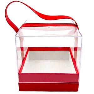 May include: A clear, square gift box with a red base and trim. The box has a red ribbon handle and a clear lid. The interior of the box is white. This decorative box is suitable for various gifts.