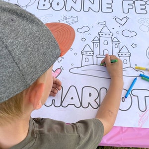 May include: A child colors a castle drawing on a white sheet of paper with the words "Bonne Fete Charlotte" at the top. The child is using colorful crayons to color the castle and the words.