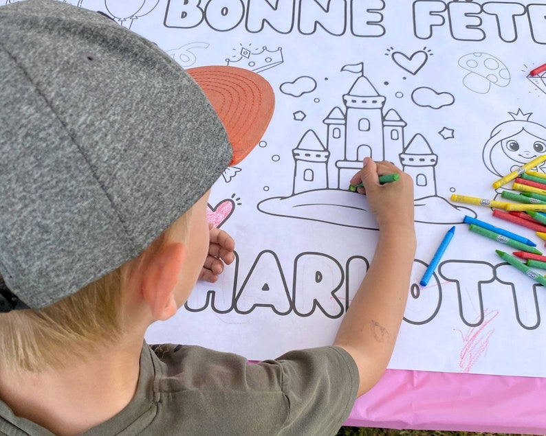 May include: A child colors a castle drawing on a white sheet of paper with the words "Bonne Fete Charlotte" at the top. The child is using colorful crayons to color the castle and the words.