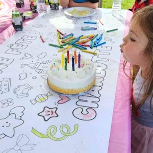 May include: A white birthday cake with colorful candles on top, sitting on a white tablecloth with a colorful drawing of a castle and the words "Une Fete Charlotte".