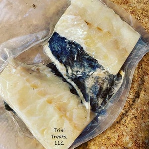 May include: Two pieces of white fish, vacuum sealed in a clear plastic bag. The fish is partially covered with a dark blue and black substance. The bag has the text "Trini Treats, LLC" printed on it.