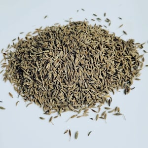 May include: A close-up image of a pile of cumin seeds. The seeds are brown and have a slightly elongated shape. They are scattered on a white surface.