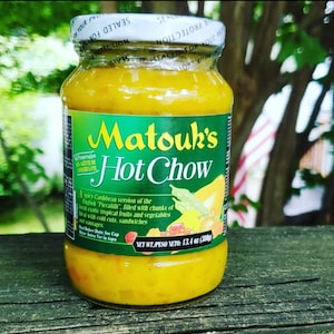 May include: A glass jar of Matouk's Hot Chow, a spicy Caribbean version of the English "Piccalilli". The jar is filled with chunks of fresh exotic tropical fruits and vegetables. The label is green and yellow with the text "Matouk's Hot Chow" in large, bold letters. The jar is sitting on a wooden surface.
