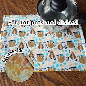 May include: A white and yellow quilted pot holder with a sloth pattern and the text "Born to be Mild". The reverse side is a yellow and orange batik pattern. The text "For hot pots and dishes!" is printed on the front.  The pot holder is shown with a silver pot on top.