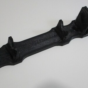 May include: A black plastic 3D printed stand with three slots for holding objects. The stand has a smooth, textured surface.