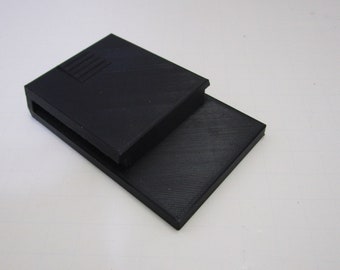 May include: A black plastic 3D printed object with a slot on one side and a raised pattern on the top.