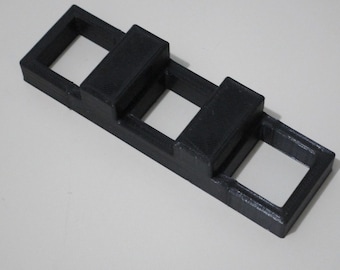 May include: A black plastic 3D printed object with three rectangular openings. The object has two raised sections on the top.