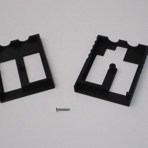 May include: Two black plastic pieces with cutouts, one with two rectangular openings and the other with a single rectangular opening and a small circular opening. A small silver screw is also visible.