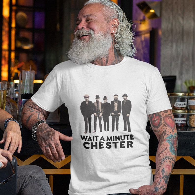 May include: White t-shirt with a black and white image of five men in suits and hats. The text "WAIT A MINUTE CHESTER" is printed below the image.