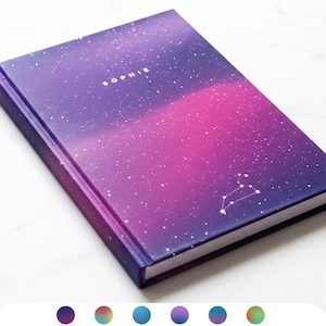 May include: A hardback notebook featuring a galaxy design in purple and pink hues. The name "SOPHIE" is printed in white on the cover, alongside a constellation. The notebook has a dark blue spine and edges.