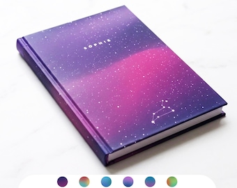 May include: A hardcover notebook with a vibrant galaxy design in shades of purple and pink. The word "SOPHIE" is printed in white on the cover, along with a constellation. The notebook has a dark blue spine and edges.