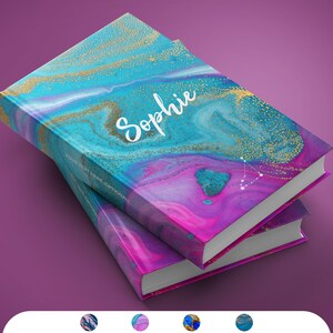 Puede incluir: Dos libros encuadernados en tapa dura apilados con una cubierta de m&aacute;rmol azul, rosa y dorado. El libro superior tiene el nombre "Sophie" escrito en cursiva blanca en la portada. El libro inferior es de un tono m&aacute;s claro de rosa y azul.