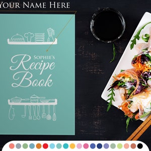 Puede incluir: Un libro de recetas turquesa con el texto "Sophie's Recipe Book" en escritura blanca. La portada presenta ilustraciones de utensilios de cocina. Un plato de rollitos de primavera y palillos chinos están sobre una superficie oscura.