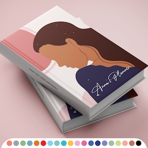 Pode incluir: Dois livros de capa dura empilhados sobre uma superfície rosa. O livro superior apresenta uma ilustração da silhueta de uma mulher com cabelo castanho comprido, um fundo rosa e branco, e o texto "Anna's Planner". A lombada diz "YOUR TEXT HERE (OPTIONAL!)".
