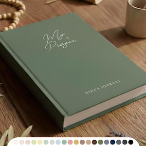 Könnte beinhalten: Ein salbeigrünes Journal mit den Worten "My Prayer" und "Nina's Journal" in weißer Schrift. Ein goldenes Kreuz, ein hölzerner Rosenkranz und eine kleine Keramiktasse sind ebenfalls auf einer Holzoberfläche zu sehen.