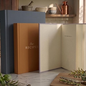 May include: Three hardcover books standing on a surface. One is a large dark blue book, one is a medium-sized orange book with the gold text Our RECIPES on the cover, and one is an open book with light-colored pages and a lavender-colored cover.