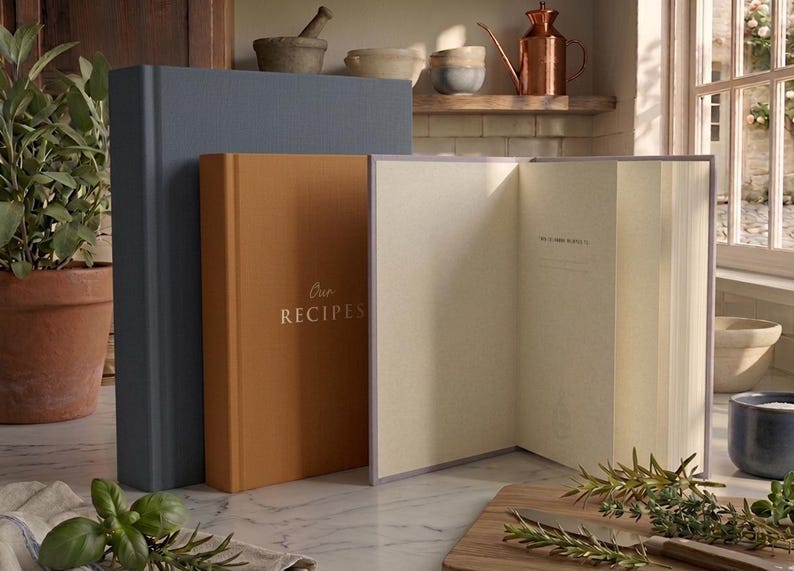 May include: Three hardcover books standing on a surface. One is a large dark blue book, one is a medium-sized orange book with the gold text Our RECIPES on the cover, and one is an open book with light-colored pages and a lavender-colored cover.