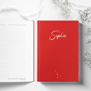 Op de afbeelding: Een rood notitieboek met de naam "Sophie" in witte letters. Het notitieboek is open en toont gelinieerde pagina's. Een sterrenbeeldontwerp staat onderaan. Witte bloemen en een lint staan op de achtergrond.
