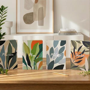 May include: Four hardcover journals with colorful abstract botanical designs. Each journal features a different name: Mariam, Nina, Melissa, and Sophie. A gold pen rests on the wooden table. Decorative plants and artwork are in the background.