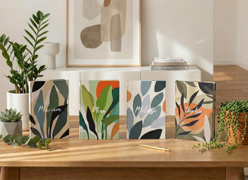 May include: Four hardcover journals with colorful abstract botanical designs. Each journal features a different name: Mariam, Nina, Melissa, and Sophie. A gold pen rests on the wooden table. Decorative plants and artwork are in the background.