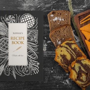 May include: A black recipe book titled "Sophie's Recipe Book" with a white and gold design, featuring illustrations of baked goods. Sliced bread and pastries are arranged on a dark surface, alongside a wooden serving board with snacks.