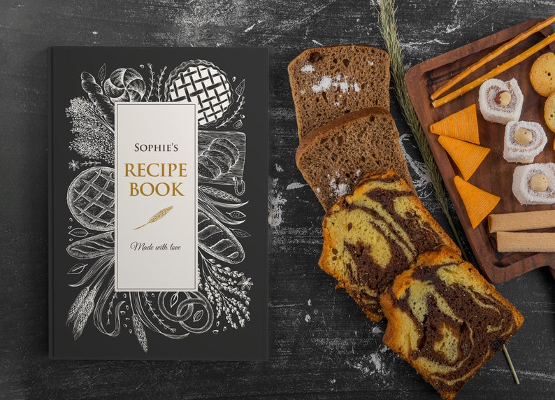 May include: A black recipe book titled "Sophie's Recipe Book" with a white and gold design, featuring illustrations of baked goods. Sliced bread and pastries are arranged on a dark surface, alongside a wooden serving board with snacks.