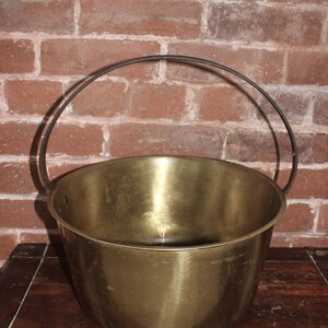 May include: A large, round, brass bucket with a metal handle. The bucket is sitting on a wooden surface.