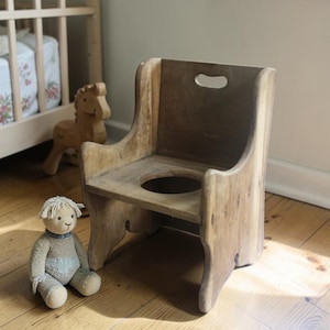 May include: A small, light brown wooden potty chair featuring a high backrest with a cutout handle, curved side armrests, and a circular opening in the seat. The chair has a natural wood grain finish and a sturdy, low-profile base design.