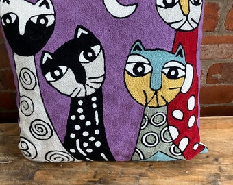 Peut inclure: Un coussin violet avec des chats brodés en noir, blanc, rouge, jaune et bleu. Les chats sont stylisés et ont de grands yeux. Il y a un croissant de lune blanc dans le coin supérieur droit.