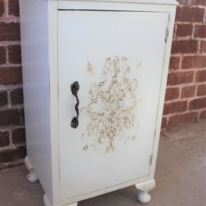 May include: A white cabinet with a single door and a decorative floral design. The cabinet has a distressed finish and a metal handle. It sits on four turned legs.