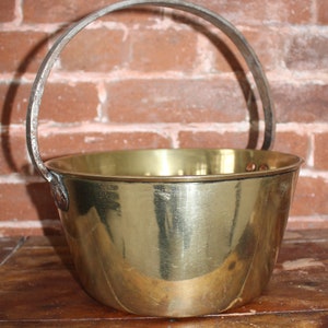 May include: A brass bucket with a metal handle. The bucket is shiny and has a rounded bottom. The bucket is sitting on a wooden surface.
