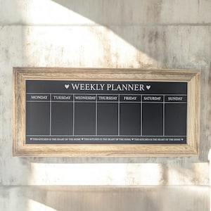 May include: A wooden framed chalkboard with a white background and black text. The chalkboard is divided into seven sections, one for each day of the week. The text at the top reads "♥ WEEKLY PLANNER ♥". The text at the bottom reads "THIS KITCHEN IS THE HEART OF THE HOME".